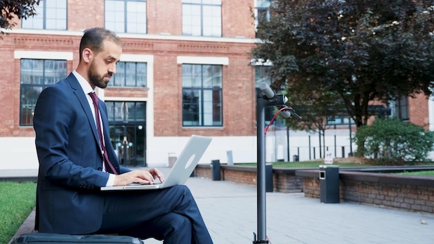 Zakenman in diplomatiek pak zittend op een bankje voor opstarten gebouw kantoor met laptopcomputer werken bij marketing presentatie typen bedrijfsstrategie. buitenconcept