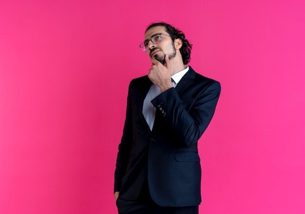 Zakenman in zwart pak en bril opzij kijken met hand op kin verbaasd staande over roze muur