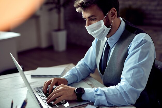 Zakenman met gezichtsmasker die op laptop op kantoor werkt tijdens een pandemie van het coronavirus