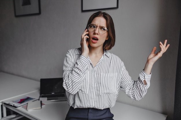 Zakenvrouw in stijlvolle blouse emotioneel praten over de telefoon. Schot van meisje met bril op achtergrond van witte tafel met briefpapier.