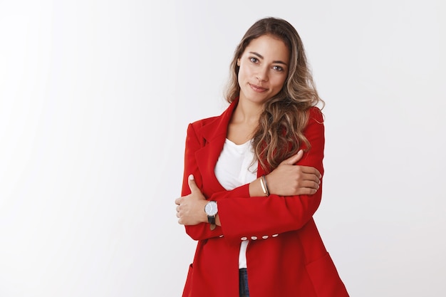 Zakenvrouw kan er zacht uitzien. studio shot aantrekkelijke vrouwelijke jonge werkende vrouw die rode jas draagt en zichzelf omhelst glimlachend schattig kantelend hoofd starend zacht, vrouwelijke werknemer die kantoorfeest bijwoont