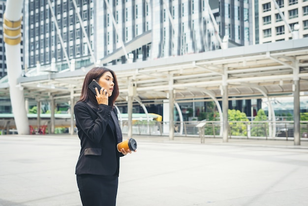 Gratis foto zakenvrouw met behulp van telefoon met koffie in de hand lopen op de straat met kantoorgebouwen op de achtergrond