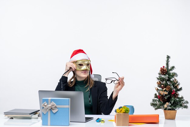 Zakenvrouw met kerstman hoed bril houden en masker zittend aan een tafel met een kerstboom en een cadeau