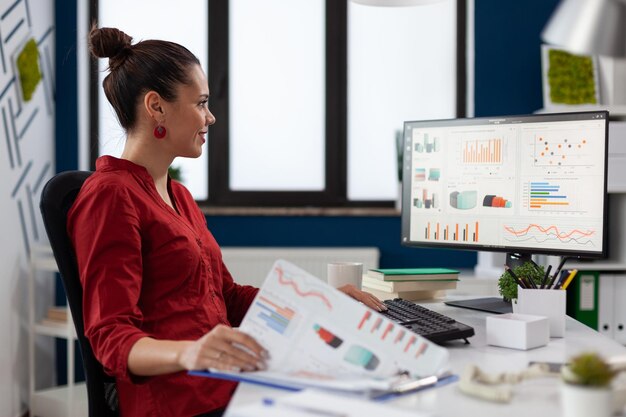 Zakenvrouw met klembord zit aan de balie in opstarten business office. Glimlachende werknemer die in rood overhemd grafieken vergelijkt. Succesvolle ondernemer die naar een desktopcomputer kijkt om bedrijfsgegevens te analyseren.