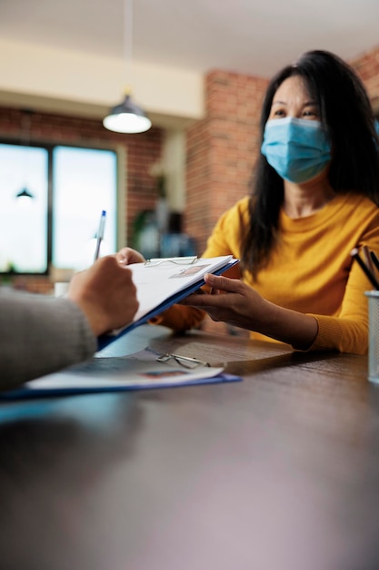 Zakenvrouw met medisch gezichtsmasker met klembord terwijl freelancer papieren ondertekent voor uitvoerend managerbaan tijdens bedrijfsinterview. team dat werkt bij zakelijke samenwerking in startbureau