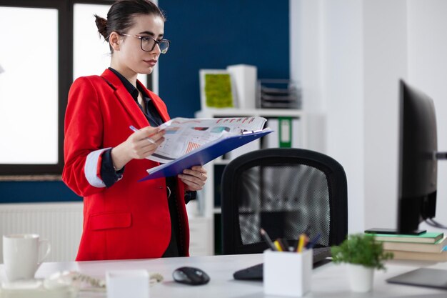 Zakenvrouw opstaan naast bureau met klembord met grafiek. Gerichte ondernemer met bril en rode jas in startbureau kijken naar bedrijfsresultaten op computerscherm.