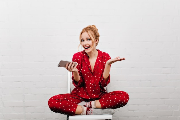 Zalig kaukasisch meisje in het gezellige nachtkostuum poseren met telefoon. portret van emotionele blonde dame in rode pyjama's met smartphone.