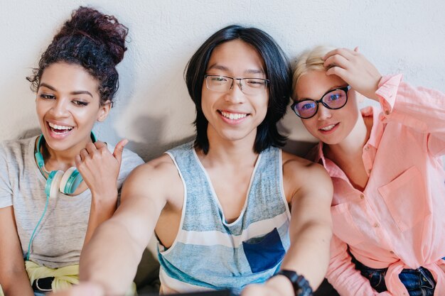 Zalig krullend Afrikaans meisje met grote koptelefoon tijd doorbrengen met haar vrienden op de universiteit. Charmante blonde vrouwelijke student poseren met glimlach terwijl haar Aziatische collega selfie maken.