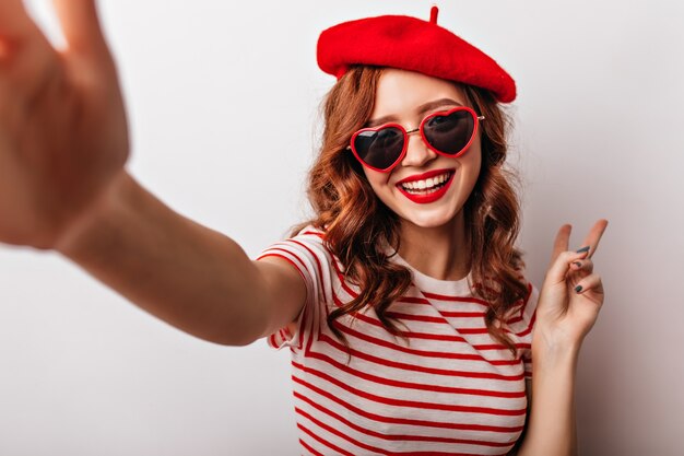 Zalige Franse vrouw in zonnebril selfie maken. Enthousiaste krullende roodharige meisje poseren op witte muur.
