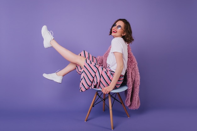 Zalige jonge dame in zonnebril gek rond tijdens fotoshoot op stoel. lachend aantrekkelijk meisje in witte schoenen.