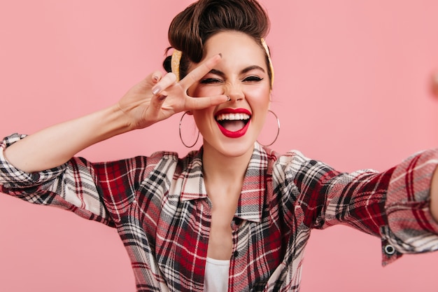 Zalige pinup meisje poseren met vredesteken op roze achtergrond. Brunette lachende dame in geruit overhemd selfie te nemen.