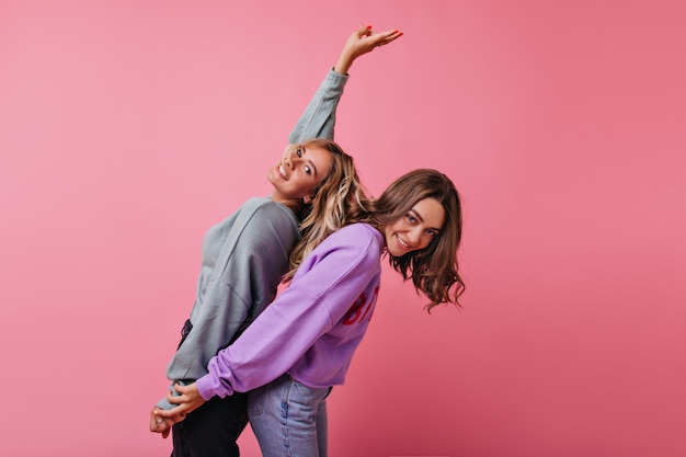 Zalige vriendinnen in straatkleren hand in hand op roze. Positieve blanke dames dansen met plezier.