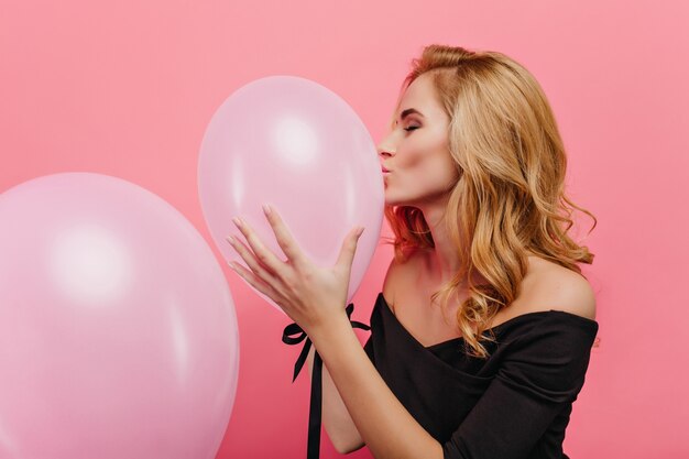 zalige vrouw met blond haar die roze ballon kust. Indoor portret van huiveringwekkende jonge dame in zwarte kledij voorbereiding voor evenement.