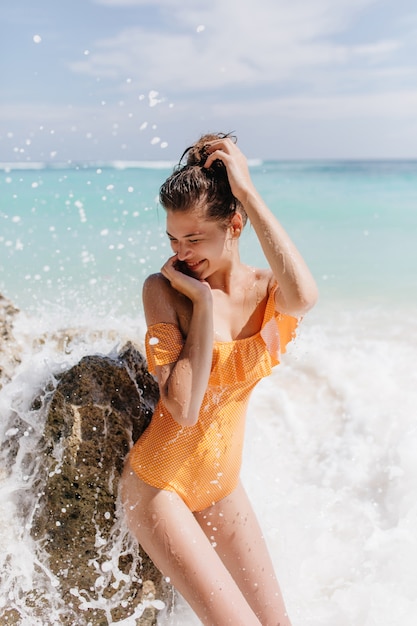 Zalige vrouw met donker haar positieve emoties uitdrukken tijdens plezier in de oceaan. Buitenfoto van mooi gelooid meisje in vintage gele zwembroek.