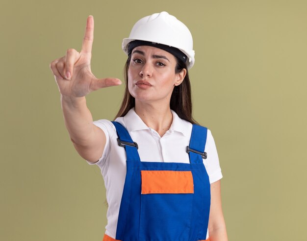 Zekere jonge bouwersvrouw in eenvormig die verliezersgebaar toont dat op olijfgroene muur wordt geïsoleerd
