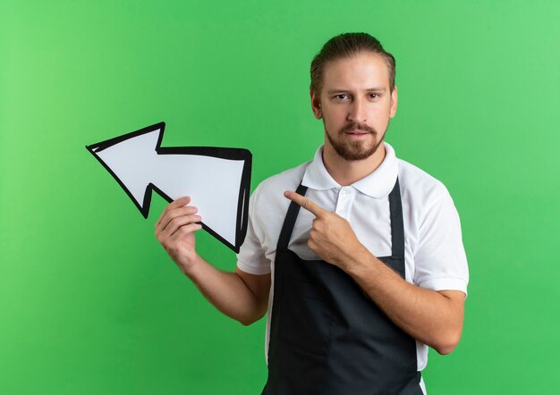 Zekere jonge knappe kapper die eenvormig het teken van de holdingspijl draagt en naar kant richt die op groen wordt geïsoleerd