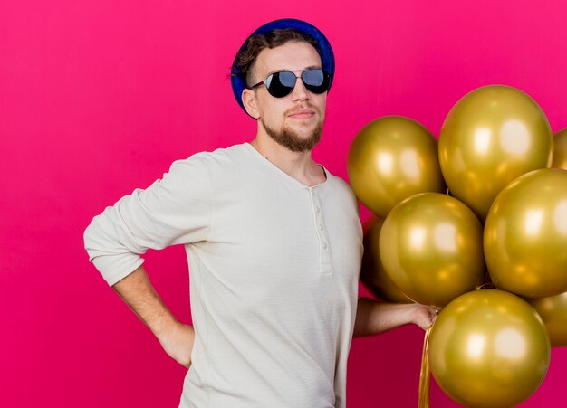 Zekere jonge knappe Slavische partijkerel die partijhoed en zonnebril draagt die ballons houdt die hand op taille houdt die voorzijde bekijkt die op roze muur wordt geïsoleerd