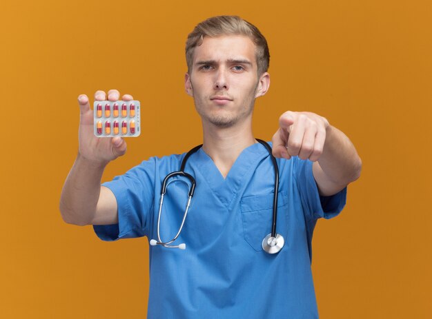 Zekere jonge mannelijke arts die artsenuniform met de pillen van de stethoscoopholding draagt die u gebaar tonen dat op oranje muur wordt geïsoleerd