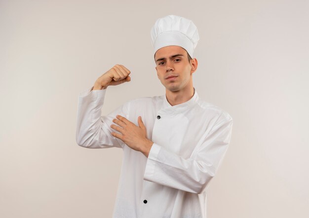 Zekere jonge mannelijke kok die eenvormige chef-kok draagt die sterk gebaar op geïsoleerde witte muur met exemplaarruimte doet