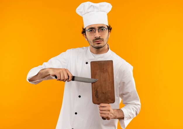 Zekere jonge mannelijke kok die eenvormige chef-kok en glazen draagt die scherpe raad en mes houden