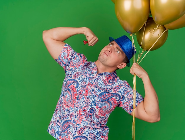 Gratis foto zekere jonge partijkerel die blauwe hoed draagt die ballons toont die sterk gebaar tonen dat op groen wordt geïsoleerd