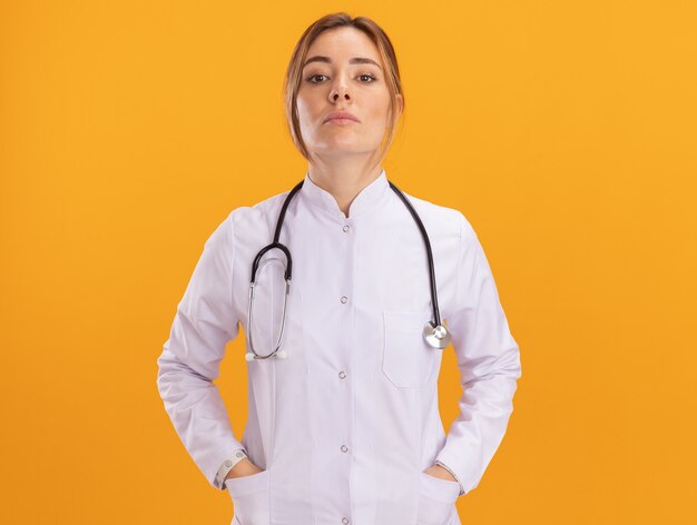 Zekere jonge vrouwelijke arts die medisch kleed met stethoscoop draagt dat handen op zak zet die op gele muur wordt geïsoleerd