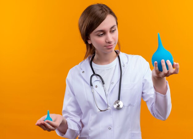 Zekere jonge vrouwelijke arts die medische mantel en stethoscoop draagt die klysma's op geïsoleerde oranje ruimte houden