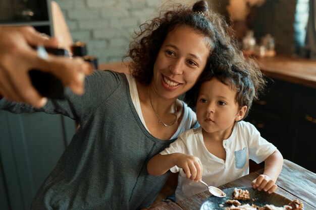 Zelf tewerkgesteld jonge vrouw die plezier heeft met haar schattige babyjongen die slimme telefoon houdt