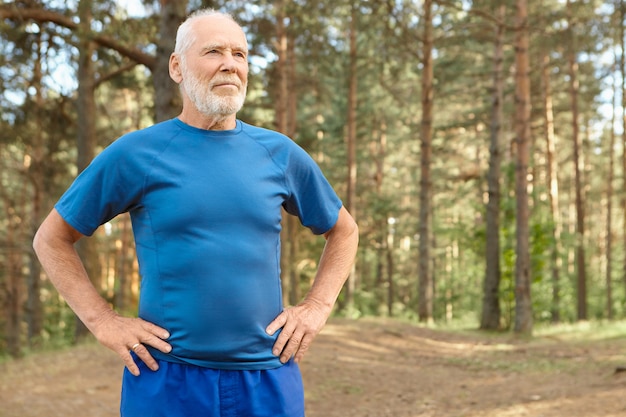 Zelfbepaalde oudere man op pensioen training buiten in dennenbos, hand in hand op zijn middel, oefeningen doen om het lichaam op te warmen voor het hardlopen. Bebaarde gepensioneerde man op adem komen na training