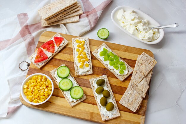 Zelfgemaakte knäckebröd toast met kwark en groene olijven, plakjes kool, tomaten, maïs, groene peper op snijplank. Gezond voedselconcept, Hoogste mening. Plat leggen