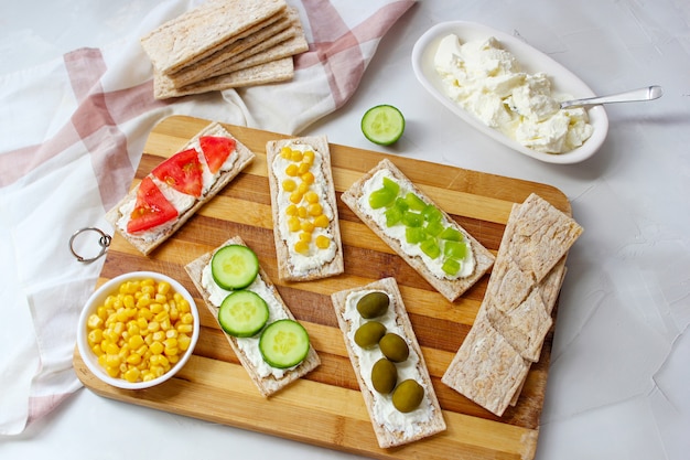 Zelfgemaakte knäckebröd toast met kwark en groene olijven, plakjes kool, tomaten, maïs, groene peper op snijplank. Gezond voedselconcept, Hoogste mening. Plat leggen