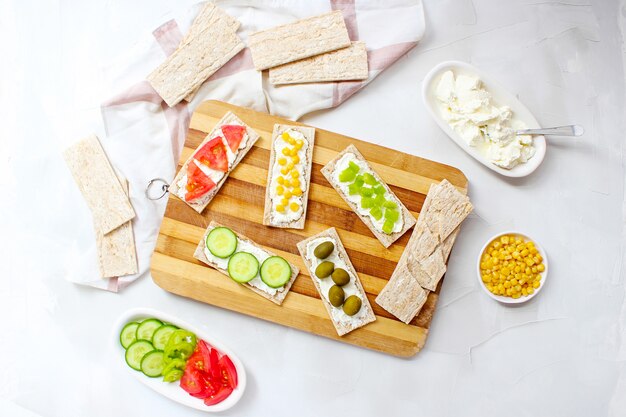 Zelfgemaakte knäckebröd toast met kwark en groene olijven, plakjes kool, tomaten, maïs, groene peper op snijplank. Gezond voedselconcept, Hoogste mening. Plat leggen