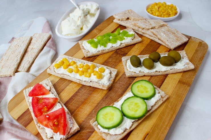 Gratis Foto zelfgemaakte knäckebröd toast met kwark en groene olijven, plakjes kool, tomaten, maïs, groene peper op snijplank. gezond voedselconcept, hoogste mening. plat leggen