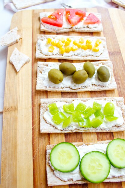 Zelfgemaakte knäckebröd toast met kwark en groene olijven, plakjes kool, tomaten, maïs, groene peper op snijplank. Gezond voedselconcept, Hoogste mening. Plat leggen