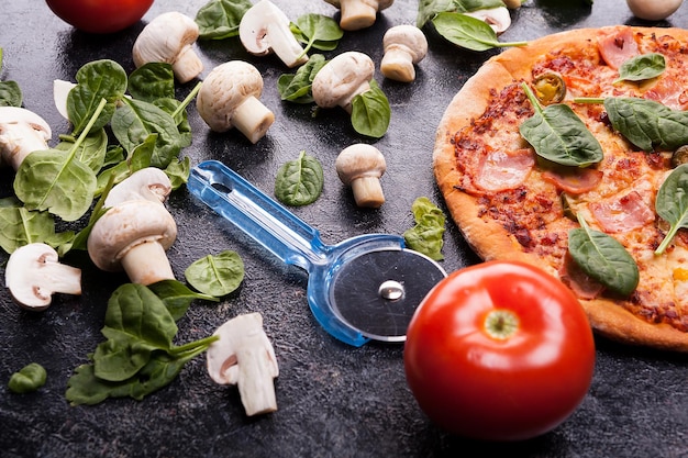 Zelfgemaakte pizza naast champignons, babyspinazie en tomaten met een pizzasnijder op donkere houten ondergrond