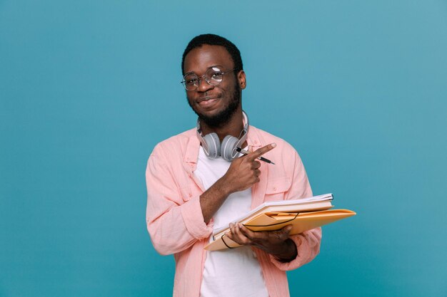 Zelfverzekerd bedrijf boeken met pen jonge Afro-Amerikaanse man met koptelefoon op nek geïsoleerd op blauwe achtergrond