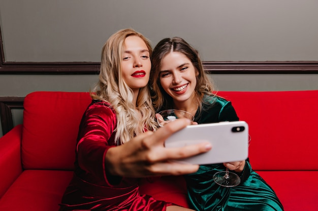 Zelfverzekerd blond meisje met telefoon voor selfie op feestje. Blanke dames champagne drinken op de bank.