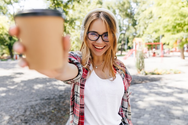 Zelfverzekerd glimlachend meisje poseren in park. vrolijke blonde dame kopje koffie houden op aard.