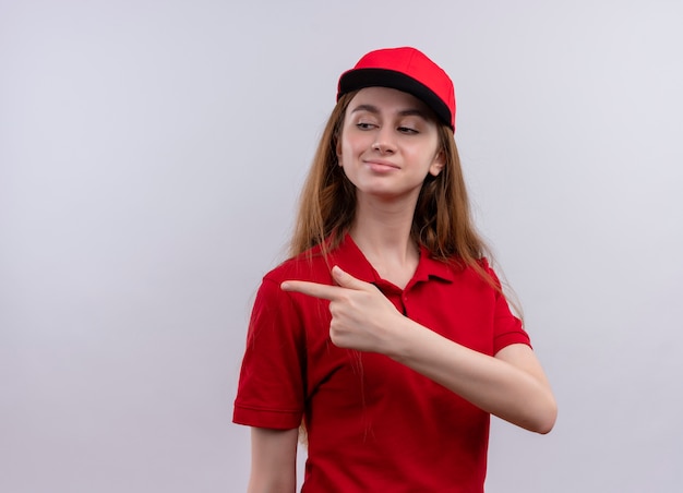 Zelfverzekerd jong bezorgmeisje in rood uniform wijzend naar de linkerkant op geïsoleerde witte ruimte met kopie ruimte