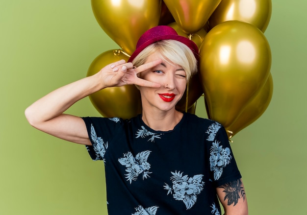 Zelfverzekerd jong blond feestmeisje met feestmuts staan voor ballonnen doen vredesteken en knipogen naar camera geïsoleerd op olijfgroene achtergrond