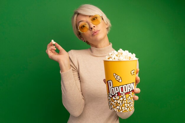 Zelfverzekerd jong blond meisje met een zonnebril die een emmer popcorn uitrekt en popcornstukken vasthoudt die op een groene muur met kopieerruimte zijn geïsoleerd