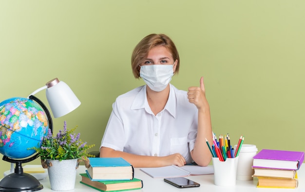 Zelfverzekerd jong blond studentenmeisje met een beschermend masker zittend aan een bureau met schoolhulpmiddelen die naar een camera kijken die duim omhoog laat zien op een olijfgroene muur