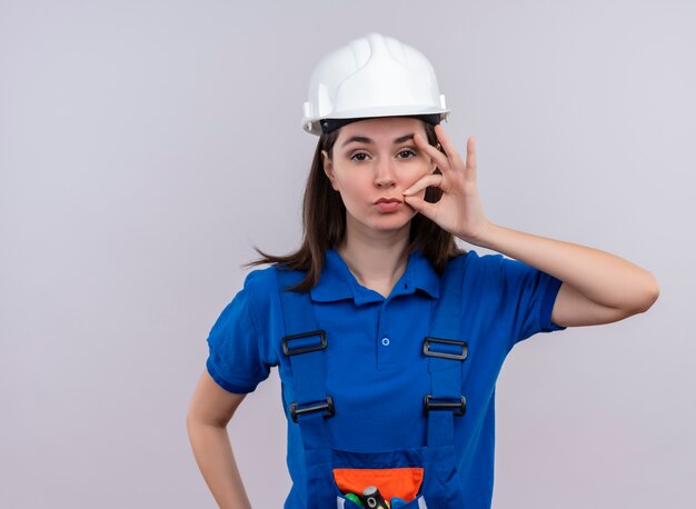 Zelfverzekerd jong bouwersmeisje met witte veiligheidshelm en blauwe uniforme ritssluitingen mond op geïsoleerde witte achtergrond