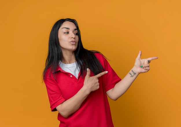 Zelfverzekerd jong donkerbruin Kaukasisch meisje dat een rood overhemd draagt kijkt en wijst naar kant met twee handen die op oranje muur worden geïsoleerd