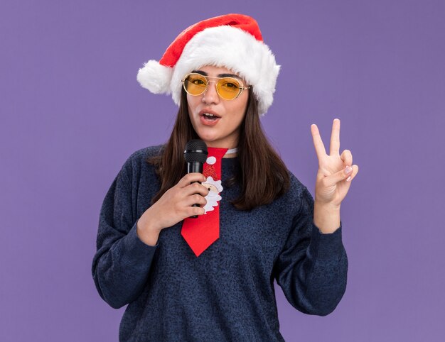 Zelfverzekerd jong Kaukasisch meisje in zonnebril met kerstmuts en santa stropdas gebaren overwinningsteken en houdt microfoon alsof ze zingen geïsoleerd op paarse muur met kopie ruimte