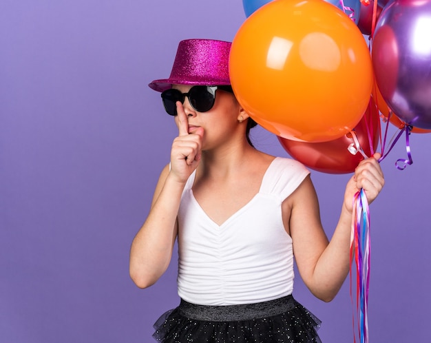 Zelfverzekerd jong Kaukasisch meisje in zonnebril met violette feestmuts die heliumballonnen vasthoudt en stiltegebaar doet geïsoleerd op paarse muur met kopieerruimte