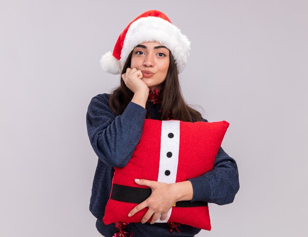 Zelfverzekerd jong Kaukasisch meisje met kerstmuts en slinger om nek houdt versierd kussen en zet vuist op kin