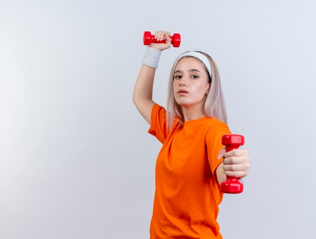 Zelfverzekerd jong Kaukasisch sportief meisje met beugels dragen hoofdband en polsbandjes staat zijwaarts met halters geïsoleerd op een witte muur met kopie ruimte