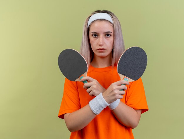 Zelfverzekerd jong Kaukasisch sportief meisje met beugels met hoofdband en polsbandjes houdt pingpongrackets vast