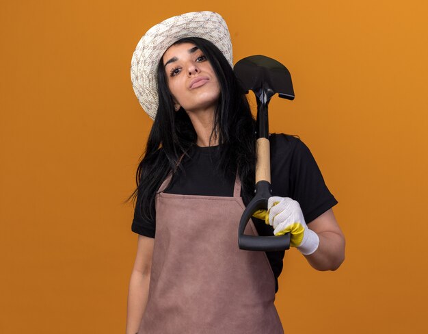 Zelfverzekerd jong Kaukasisch tuinmanmeisje met uniform en hoed met tuinmanhandschoenen met schop geïsoleerd op een oranje muur met kopieerruimte